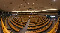 Panoramic view of the European Parliament hemicycle in Brussels, showing curved rows of empty desks and chairs, a central podium, and EU flags in the background.